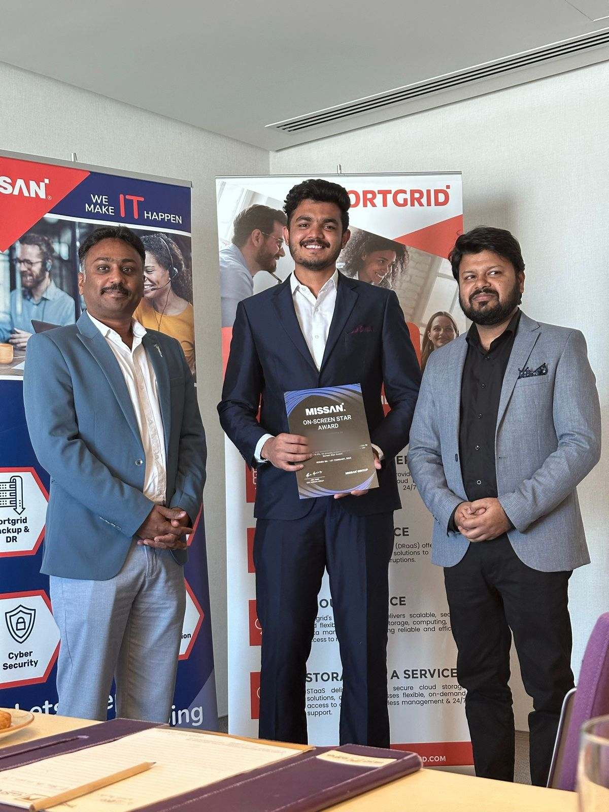 Three Missan staff in suits, the one in the middle holding an award certificate, in front of Missan and FortGrid banners