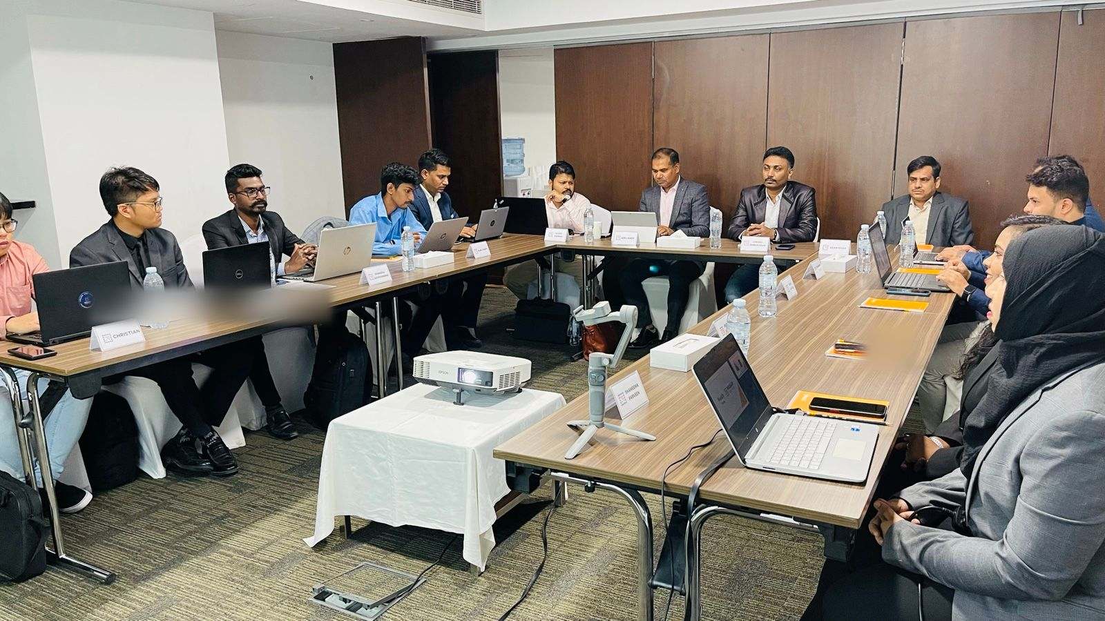 About fourteen people in business dress working on laptops around a U-shaped meeting table