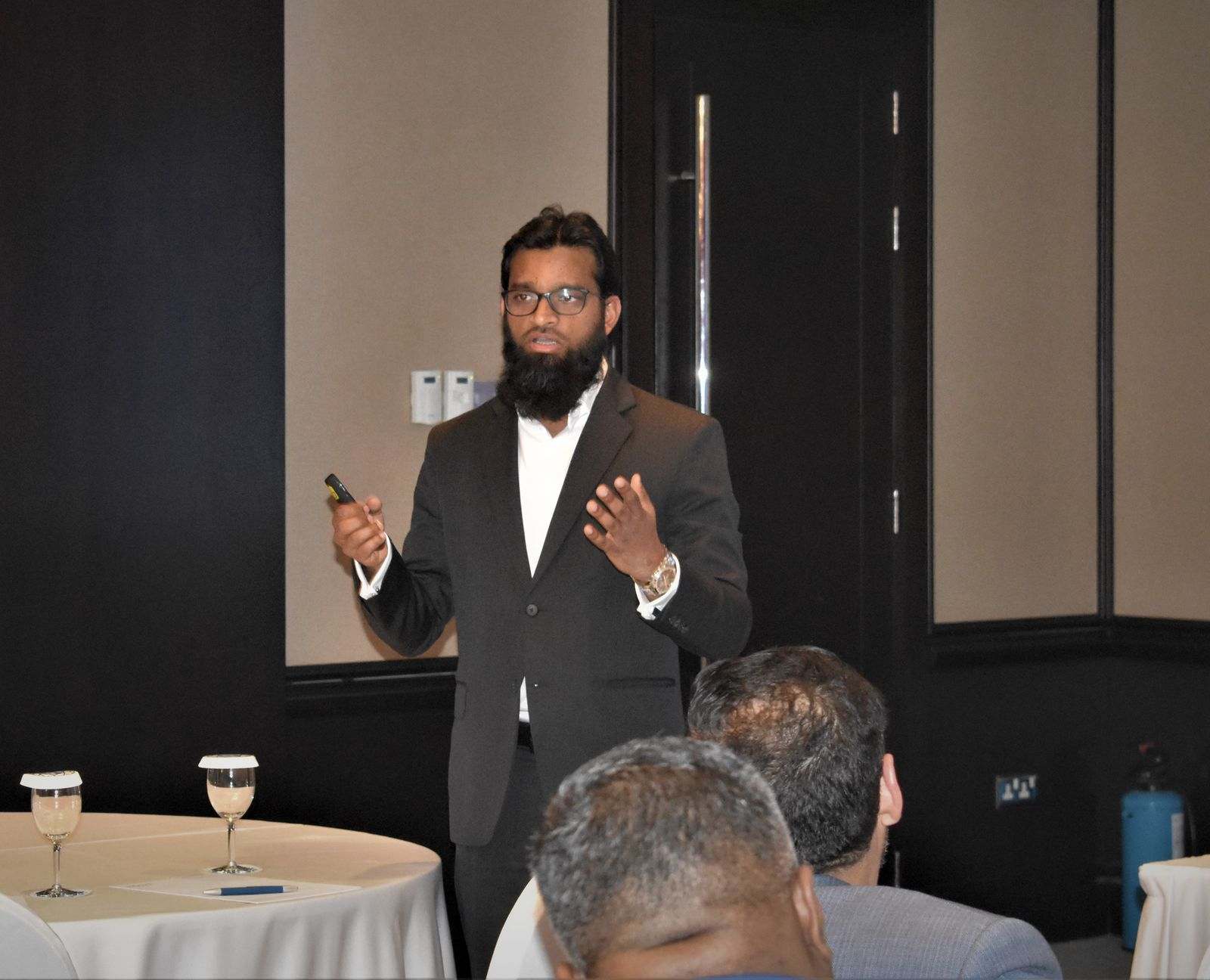 A presenter in a dark suit speaking to an audience seated at round tables