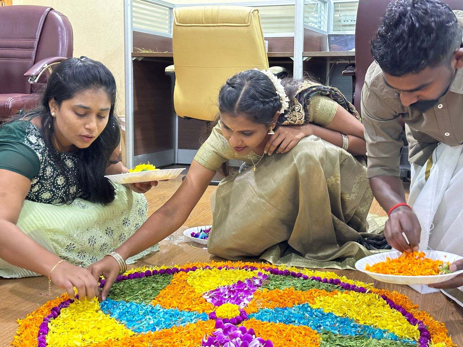 Three Missan colleagues kneeling around a colourful flower pookalam among the office desks