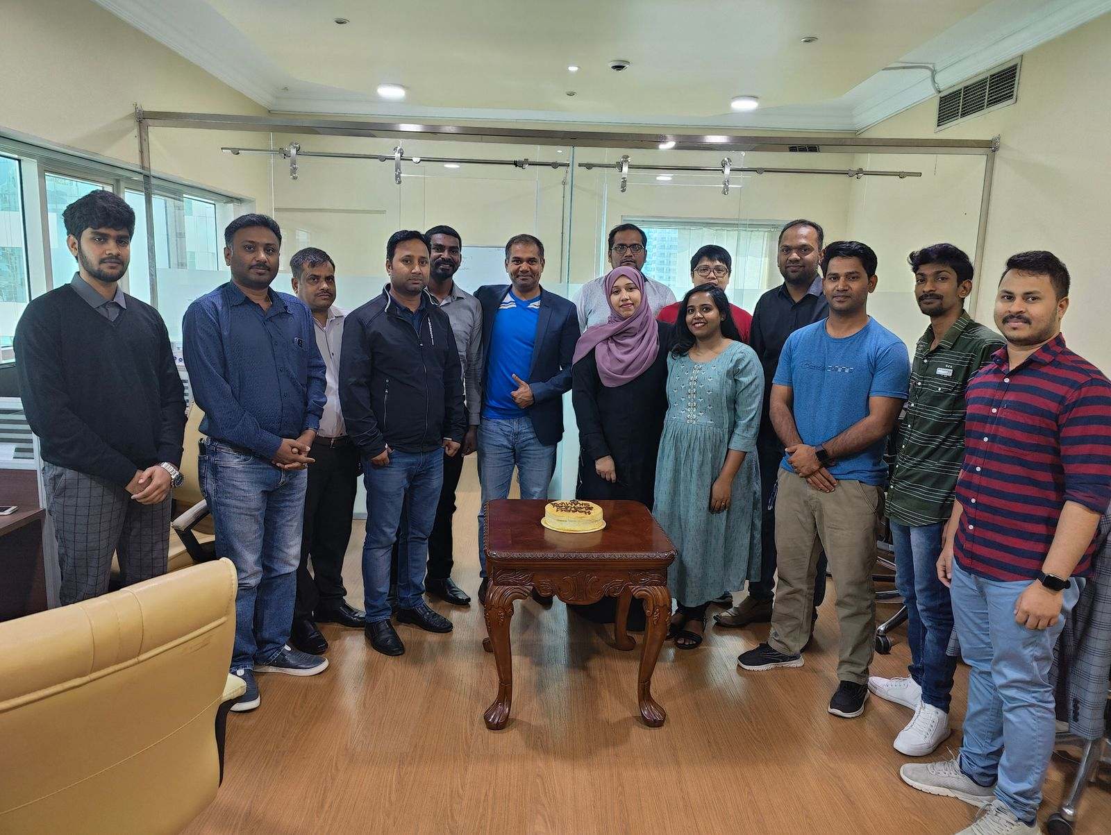 Fifteen Missan staff standing in a bright office with glass partitions, around a carved table with a cake